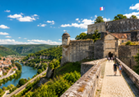 Patrimoine en Franche-Comté 6 sites incontournables à visiter cette année La Franche-Comté possède un patrimoine qui surprend souvent les visiteurs. Beaucoup connaissent la Citadelle de Besançon ou les paysages du Jura. Pourtant, la région abrite également des sites historiques parmi les plus remarquables de France. Certaines visites permettent de remonter plusieurs siècles en arrière. D'autres racontent l'histoire de personnages célèbres, d'architectes visionnaires ou de moines qui ont façonné le territoire. Bonne nouvelle, plusieurs de ces trésors restent encore relativement préservés du tourisme de masse. Si vous recherchez des idées de sorties culturelles pour cette année, ces lieux méritent clairement une place dans votre programme. La Saline Royale d'Arc-et-Senans, un monument unique en France Peu de visiteurs s'attendent à découvrir un site aussi impressionnant en arrivant à Arc-et-Senans. Pourtant, la première vision de la Saline Royale laisse rarement indifférent. Construite entre 1775 et 1779 sous l'impulsion de Claude Nicolas Ledoux, cette ancienne manufacture de sel dépasse largement sa fonction industrielle d'origine. L'architecte souhaitait créer une cité idéale où l'organisation du travail, de l'habitat et de la vie quotidienne formerait un ensemble cohérent. Près de 250 ans plus tard, le résultat continue de fasciner. L'architecture en demi-cercle imaginée par Ledoux paraît étonnamment moderne. Les bâtiments monumentaux impressionnent autant par leurs dimensions que par leur harmonie. Pourquoi la visite vaut vraiment le détour La Saline ne se limite pas à quelques bâtiments historiques. Le site accueille régulièrement des expositions temporaires, des événements culturels et un parcours permanent qui permet de comprendre le rôle essentiel du sel dans l'économie de l'époque. Les jardins méritent également votre attention. Plusieurs espaces paysagers complètent aujourd'hui la visite. Informations pratiques Adresse Place de la Saline à Arc-et-Senans Temps de visite conseillé entre 2 et 3 heures Distance depuis Besançon environ 35 kilomètres Ornans et le musée Courbet, quand les paysages prennent vie Certaines visites permettent de comprendre une œuvre. À Ornans, c'est presque l'inverse. Les paysages donnent soudain vie aux tableaux. Ce charmant village traversé par la Loue a vu naître Gustave Courbet, figure majeure du réalisme français. Le musée installé dans son ancienne demeure rassemble de nombreuses œuvres qui permettent de découvrir son parcours artistique. Mais le véritable spectacle commence souvent après la visite. Une expérience différente des autres musées En sortant du musée, vous retrouvez immédiatement les falaises, la rivière et les vallées qui ont inspiré le peintre. De nombreux visiteurs prennent alors plaisir à comparer les paysages réels avec ceux représentés dans les tableaux. Le musée Courbet et les paysages de la vallée de la Loue forment ainsi une visite particulièrement originale. Profitez-en pour flâner dans les ruelles du centre-ville. Ornans fait partie de ces villages où l'on aime prendre son temps. La Citadelle de Besançon reste un incontournable absolu Impossible de parler du patrimoine franc-comtois sans évoquer ce monument emblématique. Perchée à plus de 100 mètres au-dessus de la ville, la Citadelle de Besançon classée au patrimoine mondial de l'UNESCO domine toute la boucle du Doubs. Construite par Vauban à la demande de Louis XIV, elle constitue l'un des plus beaux exemples d'architecture militaire française. Bien plus qu'une simple forteresse La visite occupe facilement une demi-journée. Outre les remparts et les panoramas exceptionnels, plusieurs musées sont installés à l'intérieur du site. Le musée de la Résistance et de la Déportation attire chaque année de nombreux visiteurs. Le muséum d'histoire naturelle figure également parmi les plus fréquentés de la région. Le meilleur moment pour visiter Les fins de journée offrent souvent les plus belles lumières sur Besançon. Pendant l'été, certaines visites nocturnes permettent même de découvrir le monument dans une ambiance totalement différente. Le château de Joux, des siècles d'histoire à la frontière suisse À quelques kilomètres de Pontarlier, une immense silhouette de pierre domine les montagnes. Le château de Joux surveille la vallée depuis près de mille ans. Rarement un monument aura connu autant de transformations. Forteresse médiévale, château seigneurial, prison d'État puis fort militaire, il a traversé toutes les grandes périodes de l'histoire française. Une prison qui a accueilli des personnages célèbres Le château est notamment connu pour avoir emprisonné plusieurs figures historiques importantes. Parmi elles figure Mirabeau, mais aussi Toussaint Louverture, héros de l'indépendance haïtienne. Cette richesse historique donne une dimension particulière à la visite. Le château de Joux et ses mille ans d'histoire séduisent autant les passionnés d'histoire que les familles. L'abbaye de Baume-les-Messieurs, un joyau caché au cœur du Jura Le village de Baume-les-Messieurs fait déjà partie des plus beaux sites naturels de la région. Mais beaucoup de visiteurs oublient parfois de pousser les portes de son abbaye. Et c'est dommage. Fondée au IXe siècle, elle figure parmi les plus anciennes institutions religieuses du territoire. Ce qu'il ne faut pas manquer Les stalles sculptées attirent immédiatement le regard. Le retable flamand constitue également une pièce remarquable. L'ensemble dégage une atmosphère particulièrement paisible. L'abbaye de Baume-les-Messieurs et son patrimoine religieux exceptionnel méritent largement quelques heures de visite. Profitez-en également pour découvrir la reculée, les falaises et les cascades situées à proximité. Le site combine parfaitement patrimoine historique et patrimoine naturel. Les petites fortifications oubliées de la frontière jurassienne Lorsque l'on évoque Vauban, la Citadelle monopolise souvent l'attention. Pourtant, plusieurs ouvrages défensifs méritent également une visite. Le long de la frontière franco-suisse, différentes fortifications témoignent de l'importance stratégique de la région au fil des siècles. Certaines sont très connues. D'autres restent beaucoup plus discrètes. Pourquoi ces sites méritent-ils davantage d'attention Parce qu'ils permettent de mieux comprendre l'histoire du territoire. Chaque fortification raconte une période différente, avec ses conflits, ses enjeux et ses évolutions militaires. Les visites guidées proposées durant l'été rendent souvent ces découvertes beaucoup plus vivantes. Vous pouvez aussi consulter La renaissance du vinyle à Besançon disquaires, brocantes et bons plans Comment profiter au mieux du patrimoine franc-comtois Quelques astuces permettent de rendre vos visites encore plus intéressantes. Pensez aux Journées Européennes du Patrimoine Chaque année, le troisième week-end de septembre permet de découvrir gratuitement de nombreux monuments. Certains sites habituellement fermés ouvrent exceptionnellement leurs portes. Les Journées Européennes du Patrimoine permettent d'accéder à des lieux rarement ouverts au public. Utilisez les applications de découverte culturelle Plusieurs outils permettent aujourd'hui d'obtenir des informations complémentaires directement sur place. L'application Patrimoine en Balade fait partie des plus utiles pour localiser les monuments historiques autour de vous. Privilégiez les visites guidées Beaucoup de visiteurs sous-estiment leur intérêt. Pourtant, quelques explications suffisent souvent à transformer complètement une visite. Les anecdotes, les détails architecturaux et les récits historiques donnent une profondeur supplémentaire à la découverte. Vous pouvez aussi consulter Sorties en famille autour de Besançon nos meilleures idées pour le week-end Pourquoi la Franche-Comté mérite plusieurs séjours C'est probablement ce qui surprend le plus. Même les habitants de la région continuent régulièrement à découvrir de nouveaux lieux. Entre les sites classés à l'UNESCO, les villages de caractère, les abbayes anciennes, les fortifications militaires et les musées, les possibilités sont nombreuses. Chaque secteur possède sa propre identité. Chaque vallée raconte une histoire différente. Et c'est précisément cette diversité qui fait toute la richesse du patrimoine franc-comtois. Une chose est sûre que vous soyez passionné d'histoire, amateur d'architecture ou simplement curieux de découvrir de nouveaux horizons, la Franche-Comté ne manque pas de trésors à explorer.